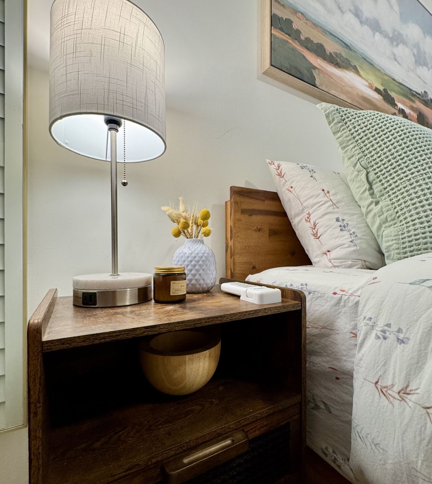 Wooden nightstand with a lamp, dried flowers and a candle beside floral bedding